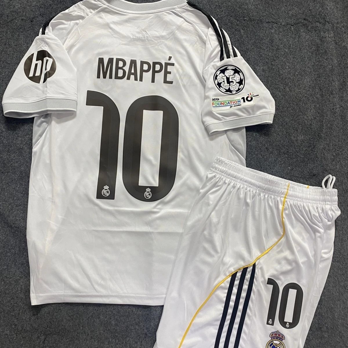 Real Madrid white short-sleeve jersey with black number 10 and name, three black stripes on sleeves, UEFA Foundation patch, matching white shorts with yellow side stripe and number